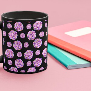 Puede incluir: Una taza de caf&eacute; negra con un patr&oacute;n de formas de cerebro rosas y moradas sobre un fondo negro.