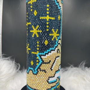 May include: A cylindrical tumbler adorned with black, yellow, and blue rhinestones. The design showcases a celestial theme with stars and a crescent moon. The tumbler is set on a black base against a fluffy, white background.