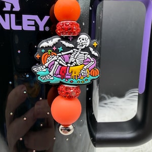 May include: A decorative charm for a tumbler featuring a central rubber skeleton figure lounging in a pool float surrounded by bats and pumpkins. The charm is accented with bright orange silicone beads and sparkling red rhinestone-encrusted spacers.