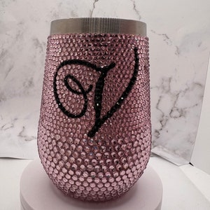 Könnte beinhalten: Ein stieloses Weinglas, das mit rosa Strasssteinen bedeckt ist. Ein großer schwarzer Buchstabe "J" ist auf dem Glas zentriert. Der obere Rand ist silberfarben. Das Glas steht auf einer weißen Oberfläche.