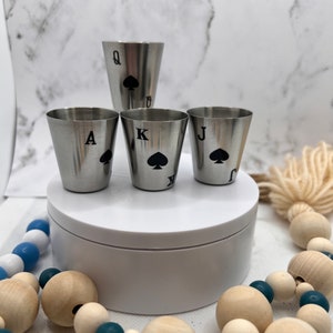 Puede incluir: Cuatro vasos de chupito de plata con símbolos de cartas de juego de picas negras. Los vasos están dispuestos en forma de pirámide con la Reina de picas en la parte superior, seguida del As, el Rey y la Sota de picas.