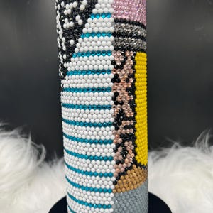 May include: A cylindrical tumbler covered in a mosaic of colorful rhinestones. The design includes sections of black and white, pink, blue and white stripes, yellow, and gray. The tumbler sits on a black base.