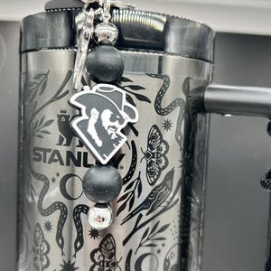 May include: A stainless steel Stanley tumbler with a black handle and a decorative charm. The charm features a black and white graphic of a man in a cowboy hat, black beads, and a silver-colored key. The tumbler is adorned with a nature-themed pattern.