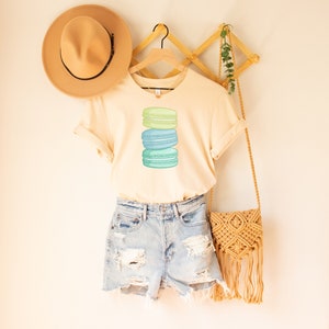 May include: A light beige t-shirt with a stack of three pastel green and blue macarons printed on the front. The t-shirt is hanging on a wooden hanger and is styled with denim shorts and a straw bag.