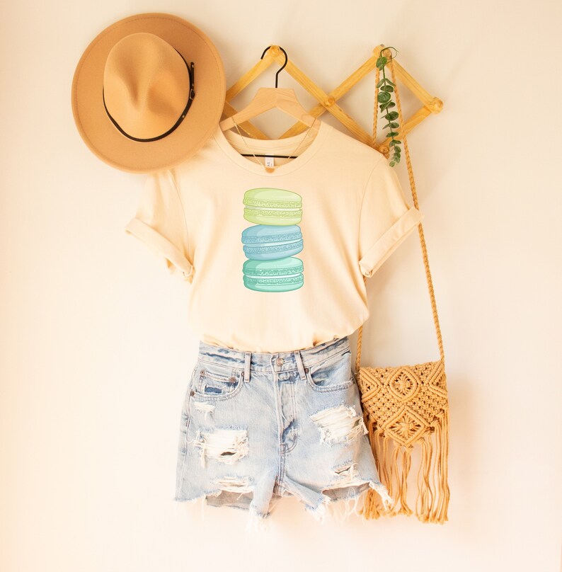May include: A light beige t-shirt with a stack of three pastel green and blue macarons printed on the front. The t-shirt is hanging on a wooden hanger and is styled with denim shorts and a straw bag.