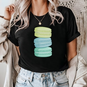 May include: A black t-shirt with a stack of three pastel macarons. The macarons are green, blue, and yellow.