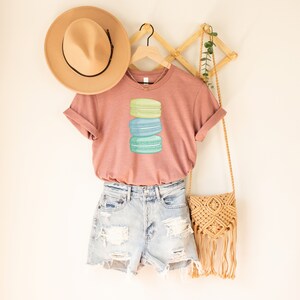 May include: A dusty rose t-shirt with a stack of three pastel macarons on the front. The macarons are green, blue, and light blue. The shirt is hanging on a wooden rack with a straw hat and a woven bag.