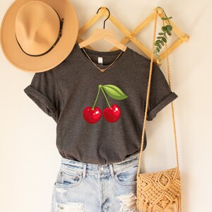 May include: Dark gray t-shirt featuring a vibrant illustration of two red cherries with a green leaf. The shirt is displayed on a wooden hanger, paired with light blue denim shorts and a woven crossbody bag. A tan hat is visible in the background.