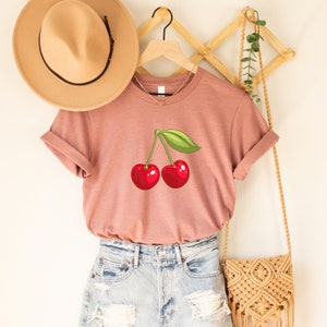 May include: A dusty rose-colored t-shirt featuring a graphic of two red cherries with a green leaf. The shirt is displayed on a wooden hanger, paired with denim shorts and a woven crossbody bag. A tan hat hangs nearby.