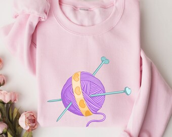 May include: A pink sweatshirt with a purple yarn ball and two knitting needles with hearts on the ends. The yarn ball has a yellow band with small hearts.