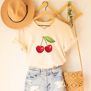 May include: Cream-colored t-shirt featuring a graphic of two red cherries with a green leaf. The shirt is displayed on a wooden hanger. A straw hat and a woven bag are also visible.