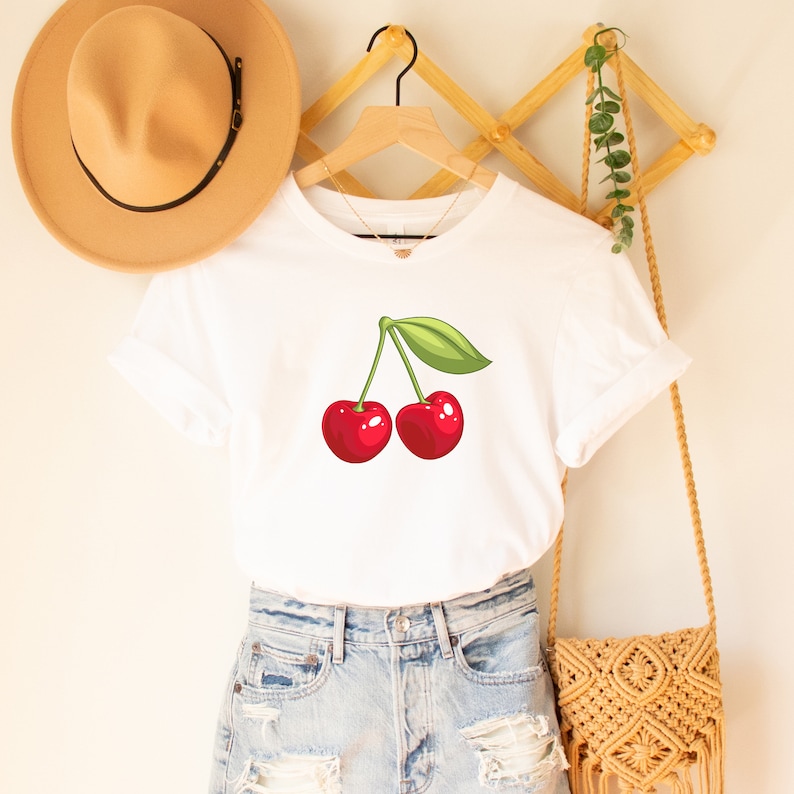 May include: White t-shirt featuring a vibrant illustration of two red cherries with a green leaf. The shirt is displayed on a wooden hanger, paired with denim shorts and a woven shoulder bag. A tan hat hangs nearby.