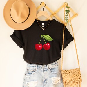 May include: Black t-shirt featuring a vibrant illustration of two red cherries with a green leaf. The shirt is displayed with a tan hat, a gold necklace, and a woven bag, suggesting a casual, summery style.