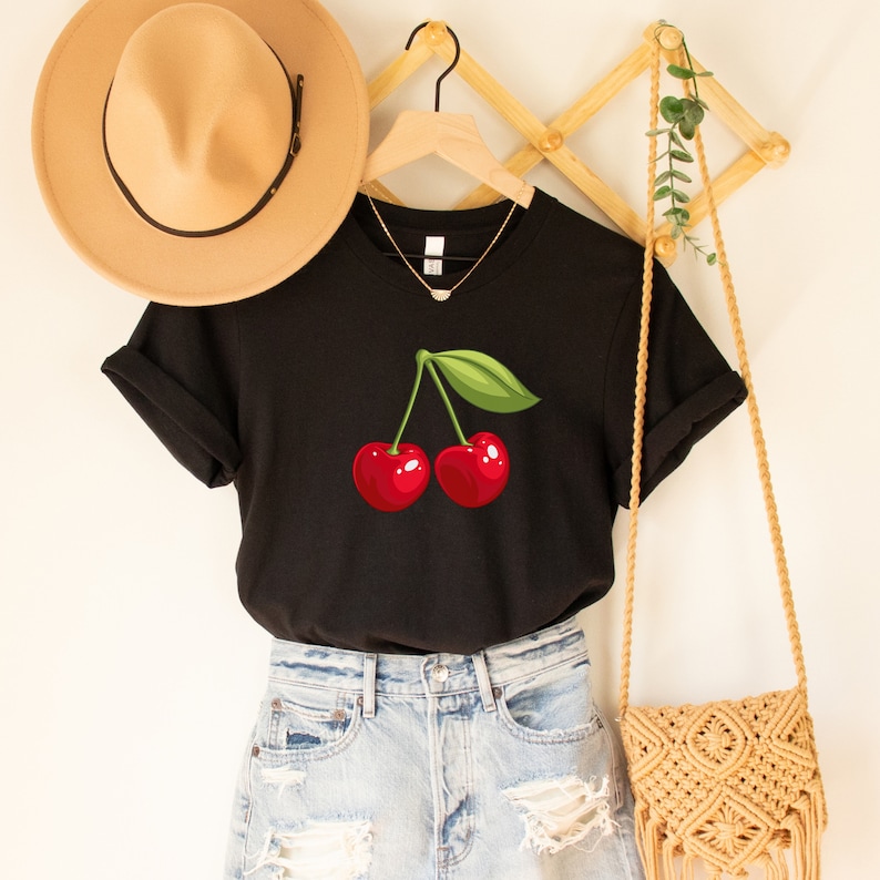 May include: Black t-shirt featuring a vibrant illustration of two red cherries with a green leaf. The shirt is displayed with a tan hat, a gold necklace, and a woven bag, suggesting a casual, summery style.