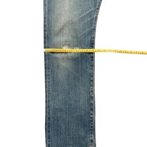 May include: Blue denim jeans with a distressed look, featuring a worn patch. A yellow measuring tape is laid across the jeans, indicating measurements. The jeans are isolated on a white background.