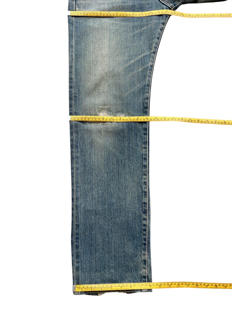 May include: Blue denim jeans with a distressed look, featuring a worn patch. A yellow measuring tape is laid across the jeans, indicating measurements. The jeans are isolated on a white background.