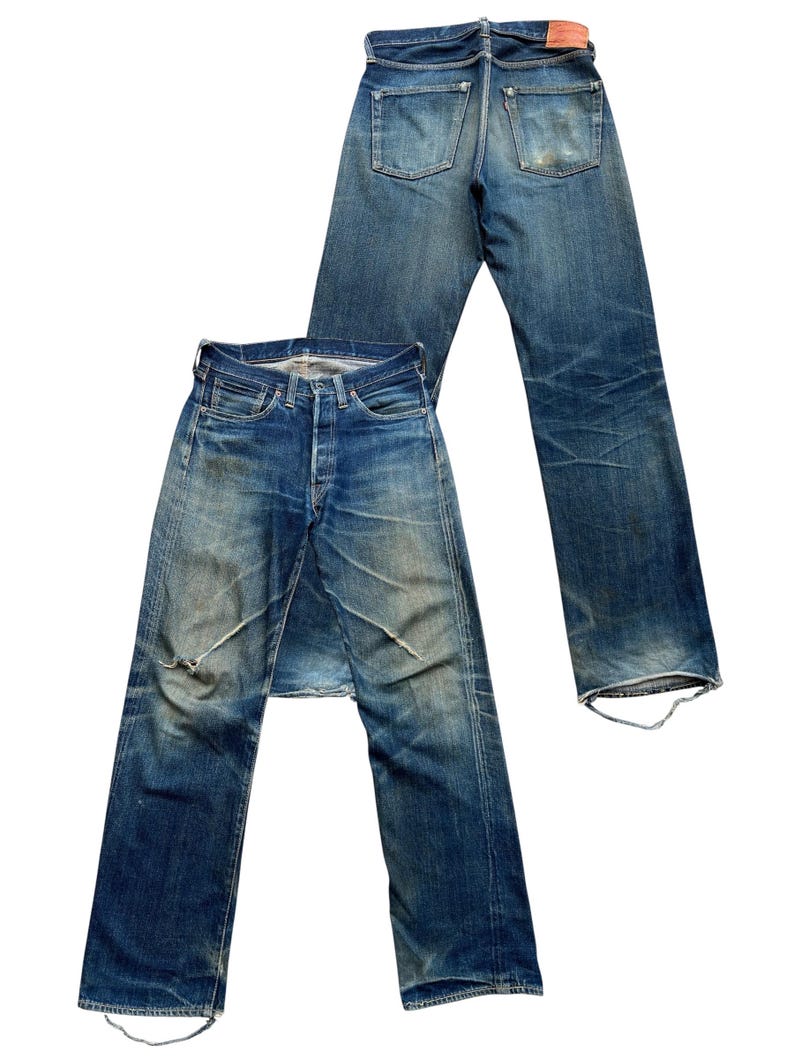 Może przedstawiać: Dwie pary dżins&oacute;w vintage z niebieskiego denimu. Jedna para leży płasko, pokazując prz&oacute;d z zapięciem na guziki i przetarciami. Druga para jest umieszczona z tyłu, pokazując tylne kieszenie i sk&oacute;rzaną naszywkę.