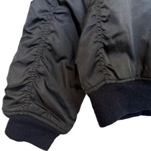 May include: Close-up of a black bomber jacket sleeve. The sleeve features gathered fabric details and a dark blue ribbed cuff. The jacket's material has a slight sheen. The bottom of the jacket has a dark blue ribbed hem with a small orange tag.