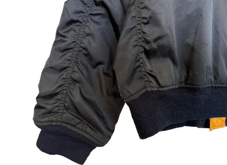 May include: Close-up of a black bomber jacket sleeve. The sleeve features gathered fabric details and a dark blue ribbed cuff. The jacket's material has a slight sheen. The bottom of the jacket has a dark blue ribbed hem with a small orange tag.