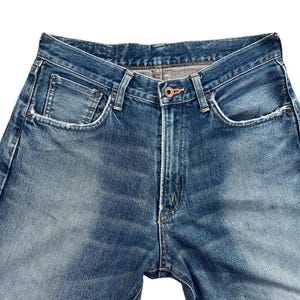 Puede incluir: Primer plano de unos vaqueros de mezclilla azules. Los vaqueros tienen un dise&ntilde;o cl&aacute;sico de cinco bolsillos, cierre de bot&oacute;n y cremallera, y un lavado descolorido. La mezclilla es de color azul medio con zonas m&aacute;s claras.