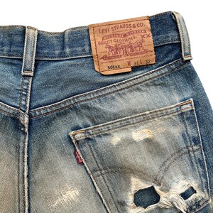 May include: Close-up of vintage blue denim jeans with a distressed pocket. The jeans feature a worn leather patch with the Levi Strauss & Co. logo and text. The jeans are size W31 L32.