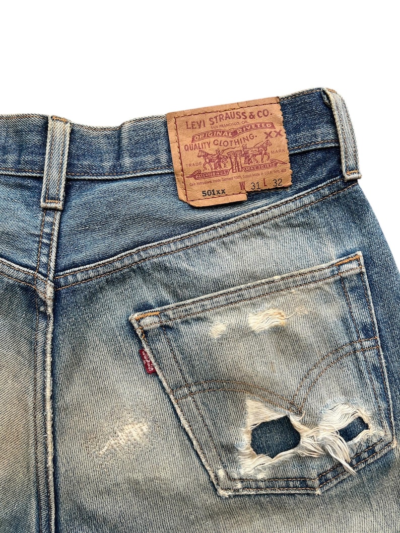 May include: Close-up of vintage blue denim jeans with a distressed pocket. The jeans feature a worn leather patch with the Levi Strauss & Co. logo and text. The jeans are size W31 L32.