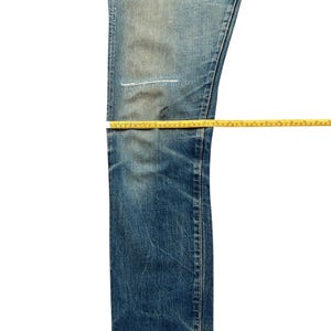 May include: Blue denim jeans with a distressed look, featuring a patched area on the leg. Yellow measuring tapes are laid across the jeans to measure the length. The jeans have a faded wash and a classic five-pocket design.