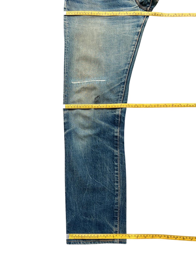 May include: Blue denim jeans with a distressed look, featuring a patched area on the leg. Yellow measuring tapes are laid across the jeans to measure the length. The jeans have a faded wash and a classic five-pocket design.