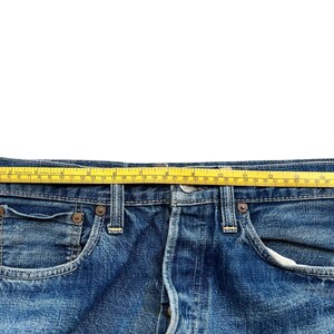 以下が含まれることがあります： 青いデニムジーンズのクローズアップ。黄色のメジャーがウエストバンドにかけられています。ジーンズはクラシックな5ポケットスタイルで、リベットとボタンフライが見えます。テープはセンチメートルで測定します。