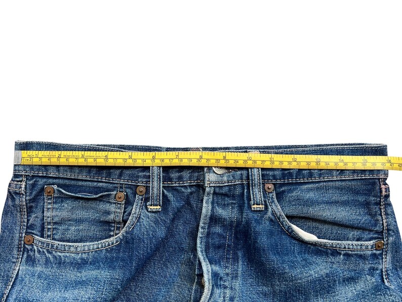 以下が含まれることがあります： 青いデニムジーンズのクローズアップ。黄色のメジャーがウエストバンドにかけられています。ジーンズはクラシックな5ポケットスタイルで、リベットとボタンフライが見えます。テープはセンチメートルで測定します。