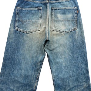 May include: Back view of blue denim jeans with two back pockets and a brown leather patch. The jeans have a faded wash and a straight leg design. The denim is a medium blue color.