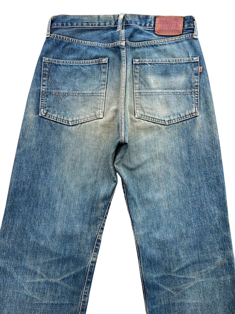 May include: Back view of blue denim jeans with two back pockets and a brown leather patch. The jeans have a faded wash and a straight leg design. The denim is a medium blue color.