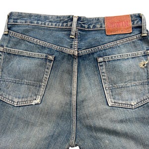 May include: Faded blue denim jeans with two back pockets and a leather patch. The jeans have a distressed look with worn areas and a small tear on one pocket. The back of the jeans is visible.