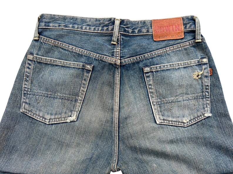 May include: Faded blue denim jeans with two back pockets and a leather patch. The jeans have a distressed look with worn areas and a small tear on one pocket. The back of the jeans is visible.