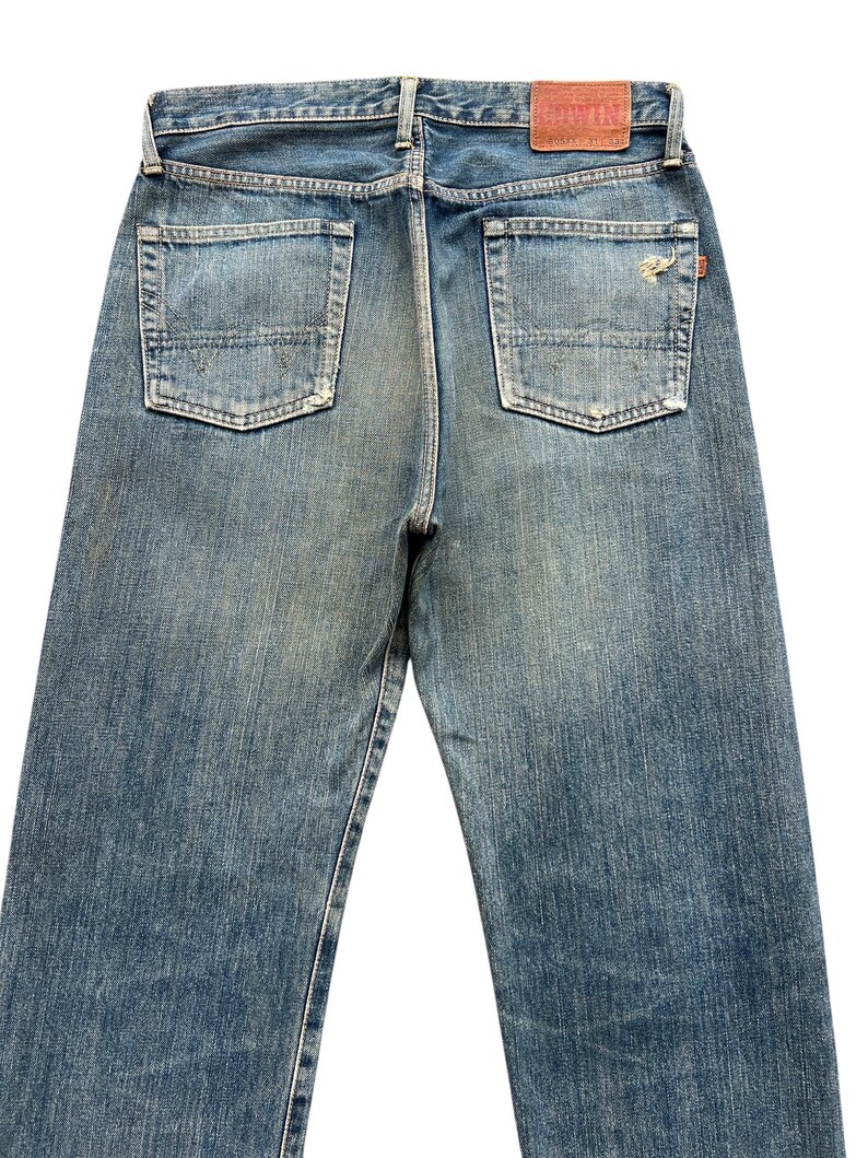 May include: Faded blue denim jeans with two back pockets and a leather patch. The jeans have a classic five-pocket design and a straight leg fit. The denim has a worn, distressed look.