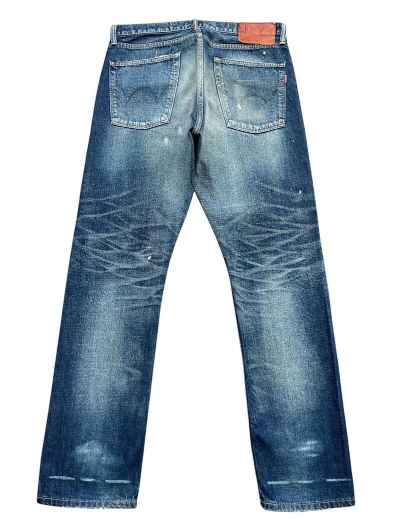 Puede incluir: Vaqueros de mezclilla azul descolorido con aspecto desgastado. Los vaqueros presentan dos bolsillos traseros, un parche de cuero y detalles desgastados. La mezclilla tiene un aspecto vintage con patrones de desgaste visibles y acentos de color claro.