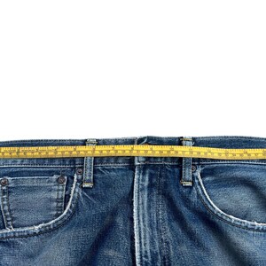 Puede incluir: Primer plano de unos vaqueros azules con una cinta m&eacute;trica amarilla sobre la cintura. Los vaqueros tienen un dise&ntilde;o cl&aacute;sico de cinco bolsillos con costuras y remaches met&aacute;licos visibles. La mezclilla tiene un aspecto descolorido y desgastado.