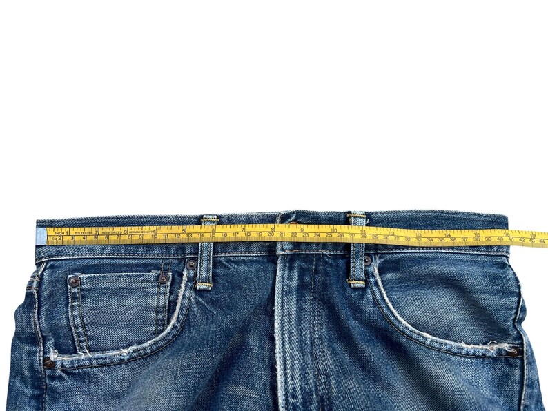 Puede incluir: Primer plano de unos vaqueros azules con una cinta m&eacute;trica amarilla sobre la cintura. Los vaqueros tienen un dise&ntilde;o cl&aacute;sico de cinco bolsillos con costuras y remaches met&aacute;licos visibles. La mezclilla tiene un aspecto descolorido y desgastado.