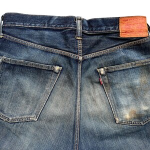 Może przedstawiać: Zbliżenie na tył niebieskich dżins&oacute;w. Dżinsy mają dwie tylne kieszenie, sk&oacute;rzaną naszywkę i czerwoną metkę. Denim ma wyblakły, zużyty wygląd.