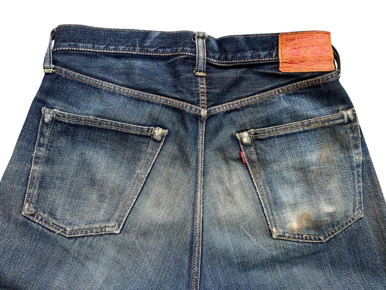 Może przedstawiać: Zbliżenie na tył niebieskich dżins&oacute;w. Dżinsy mają dwie tylne kieszenie, sk&oacute;rzaną naszywkę i czerwoną metkę. Denim ma wyblakły, zużyty wygląd.