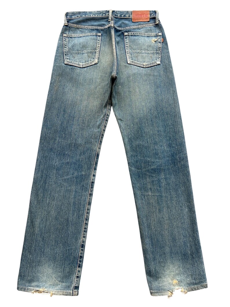 May include: Faded blue denim jeans with a classic five-pocket design. The jeans show signs of wear, including distressed areas at the hem and fading throughout. A brown leather patch is visible at the back waistband.