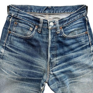 Pu&ograve; includere: Primo piano di jeans di denim blu con chiusura a bottoni, passanti per cintura e tasche anteriori e posteriori. I jeans hanno un aspetto sbiadito e usurato con segni di usura visibili e un classico design a cinque tasche.