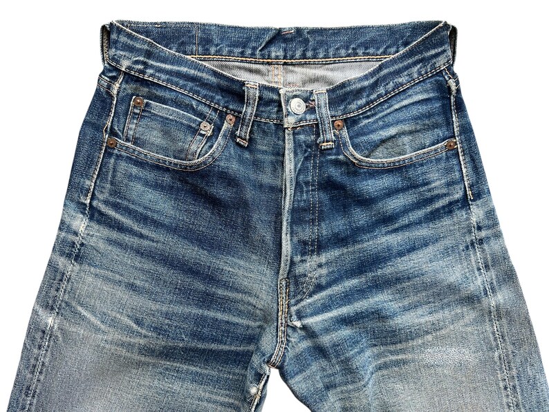Pu&ograve; includere: Primo piano di jeans di denim blu con chiusura a bottoni, passanti per cintura e tasche anteriori e posteriori. I jeans hanno un aspetto sbiadito e usurato con segni di usura visibili e un classico design a cinque tasche.