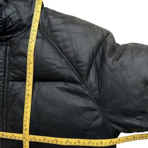 May include: Black leather jacket with a quilted design. A yellow measuring tape is draped across the jacket, showing measurements in inches. The jacket has a high collar and a padded design.