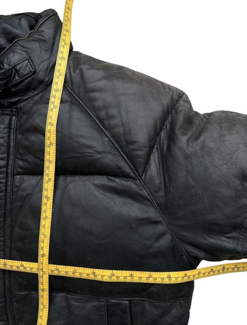 May include: Black leather jacket with a quilted design. A yellow measuring tape is draped across the jacket, showing measurements in inches. The jacket has a high collar and a padded design.