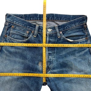 以下が含まれることがあります： ボタンフライ、ポケット、ベルトループが付いた青いデニムジーンズのクローズアップ。 黄色のメジャーがジーンズの上に置かれ、測定値を示しています。 ジーンズは色あせ、使い古された外観をしています。