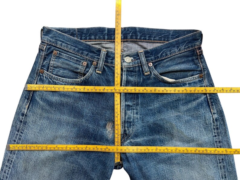 以下が含まれることがあります： ボタンフライ、ポケット、ベルトループが付いた青いデニムジーンズのクローズアップ。 黄色のメジャーがジーンズの上に置かれ、測定値を示しています。 ジーンズは色あせ、使い古された外観をしています。