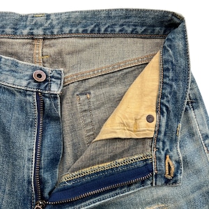 May include: Close-up of blue denim jeans with a partially unzipped fly. The jeans show signs of wear and tear, with visible fading and distressing. The zipper is silver, and the button is a dark metal. The interior lining is a light tan color.
