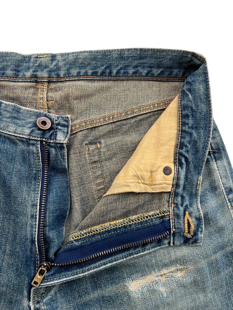 May include: Close-up of blue denim jeans with a partially unzipped fly. The jeans show signs of wear and tear, with visible fading and distressing. The zipper is silver, and the button is a dark metal. The interior lining is a light tan color.