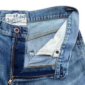 Pu&ograve; includere: Primo piano di jeans blu in denim con una cerniera in ottone parzialmente aperta. I jeans hanno una chiusura con bottone e una piccola etichetta con la scritta "New Vintage". Il denim ha un aspetto sbiadito.